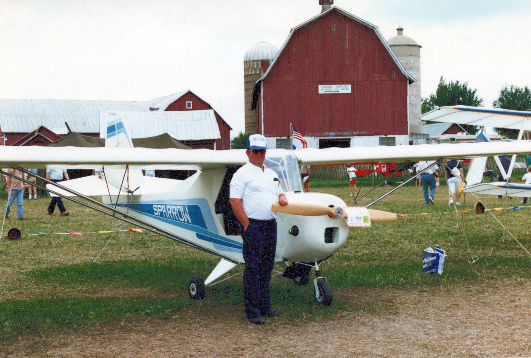 Man standing beside a white and blue Carlson Sparrow at a fly-in, with a red barn behind