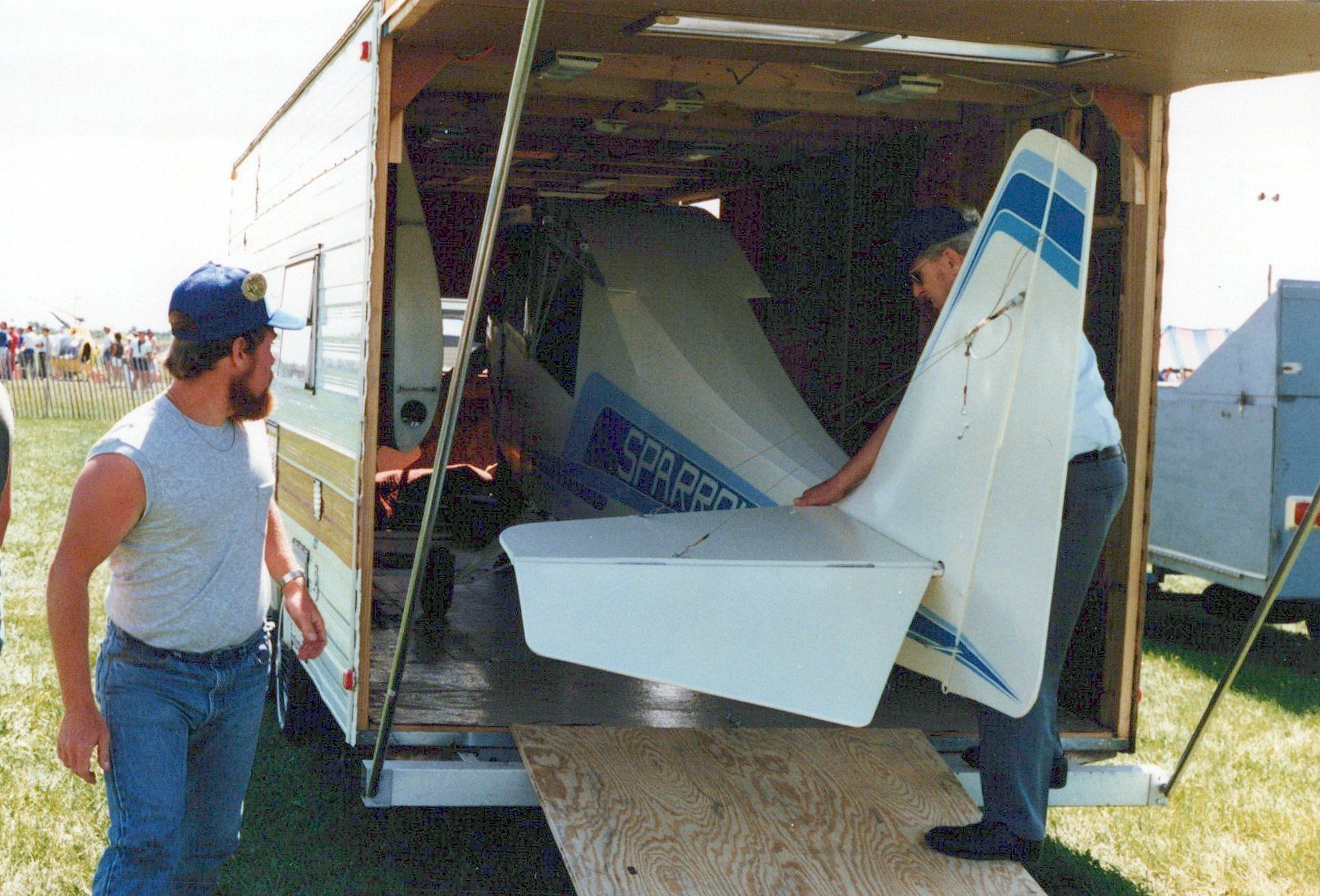 Two men loading the fuselage of a Carlson Sparrow into an enclosed trailer at a fly-in