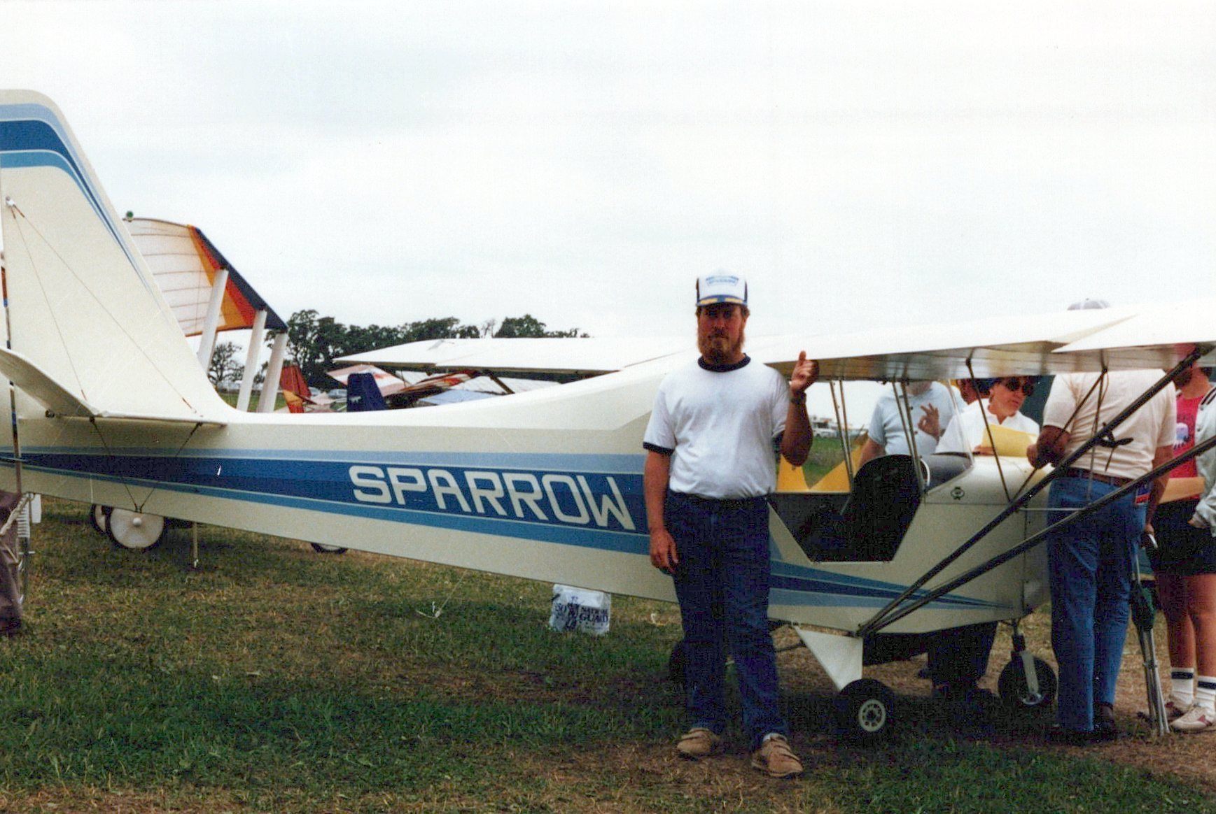 Man standing beside a white and blue Carlson Sparrow with Sparrow lettering at an airshow